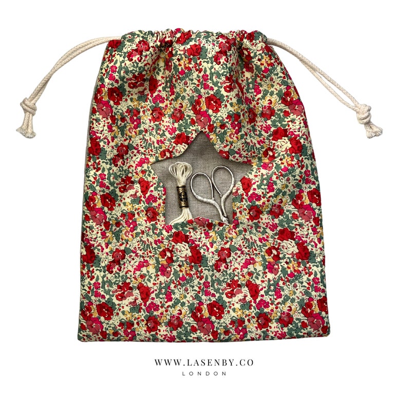May include: A white drawstring bag with a red and green floral print. The bag has a star-shaped opening with a white thread and scissors inside. The bag has the text "www.lasenby.co LONDON" printed on the bottom.