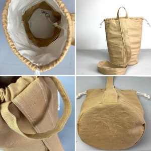 May include: Tan fabric bucket bag with a drawstring closure and a shoulder strap. The bag has a white interior lining and a small interior pocket with a metal clasp. The bag has a handle on top and a rounded bottom.