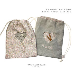 May include: Sewing pattern for a sustainable gift bag. Two fabric bags are shown, one with a floral print and the other a solid grey. Both bags have a heart-shaped opening and a drawstring closure. The grey bag has the word "Sara" embroidered on it.