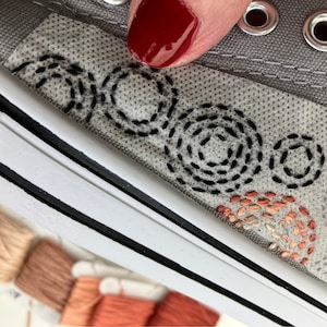 Peut inclure: Gros plan d'une chaussure en toile grise avec des détails brodés. Des cercles cousus noirs et des demi-cercles cousus orange et pêche ornent le côté de la chaussure. Semelle blanche avec bordure noire et œillets métalliques.