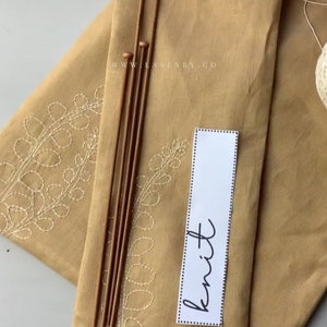 Könnte beinhalten: Ein beigefarbener Stoff mit einem weißen gestickten Blattdesign. Der Stoff ist gefaltet und hat oben Holzstricknadeln und eine weiße Wollknäuel. Eine weiße Karte mit dem Wort "stricken" in Schwarz ist auf dem Stoff.