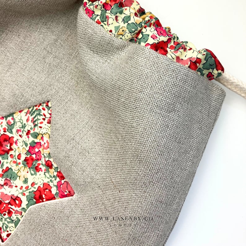 May include: A beige drawstring bag with a star-shaped patch made of floral fabric. The bag has a white drawstring and the text "www.lasenbyco.com" is printed on the bottom.