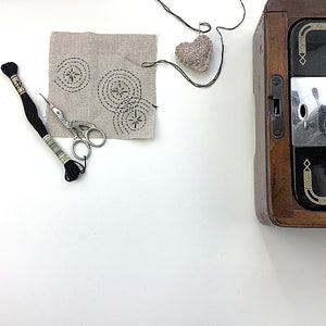 May include: A close-up of a beige fabric square with black embroidery of three circular designs. A pair of silver embroidery scissors, black thread, and a small heart-shaped pincushion are also visible.