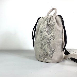 Pode incluir: Uma bolsa de cordão bege com alças pretas e um padrão de círculos pretos bordados na frente.
