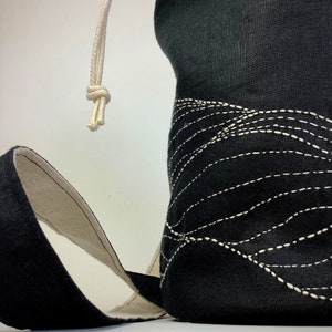 May include: Black fabric bag with a white drawstring closure and a cream-colored strap. The bag features a decorative wave-like pattern created with white stitching. The interior of the strap is cream-colored.