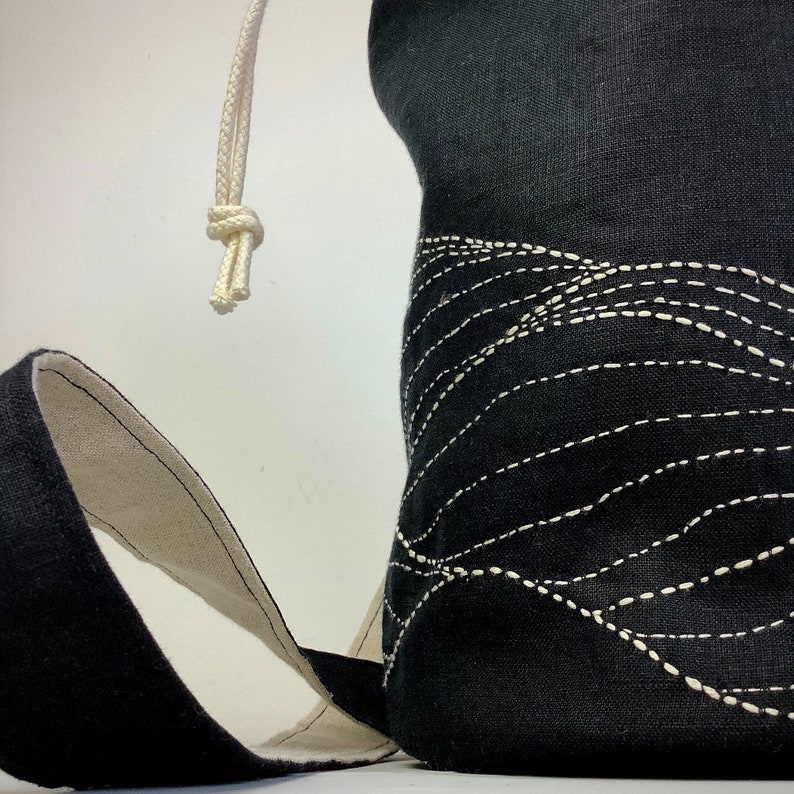 May include: Black fabric bag with a white drawstring closure and a cream-colored strap. The bag features a decorative wave-like pattern created with white stitching. The interior of the strap is cream-colored.