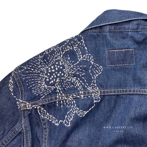 May include: Close-up of a dark blue denim jacket with a white floral embroidery design on the upper back. The jacket has a classic collar and a small rectangular patch. The text "WWW.LASENBY.CO LONDON" is visible.