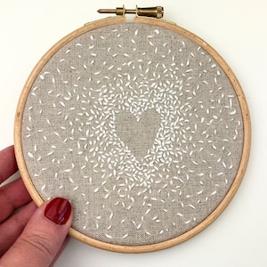 May include: An embroidered heart design in a wooden hoop. The heart shape is formed by the negative space, surrounded by white stitches on a beige fabric. The hoop is secured with a brass screw.