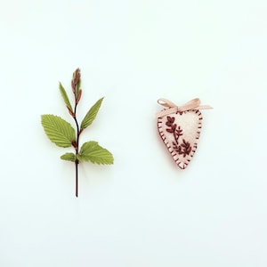 May include: A small, heart-shaped ornament made of cream-colored felt, featuring dark brown embroidered floral details and a light pink ribbon bow at the top. Beside it lies a single green stem with several serrated leaves and small buds.