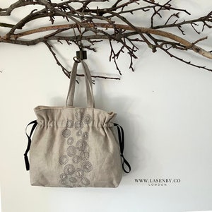 May include: A beige linen tote bag with black drawstring closures and a black embroidered design of circles. The bag is hanging from a hook on a white wall.