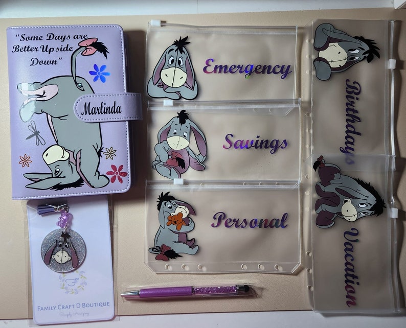 May include: A light purple A6 binder featuring Eeyore, the text "Some Days are Better Up Side Down" and "Marlinda", five clear zipper pouches with Eeyore graphics and labels for Emergency, Savings, Personal, Birthdays, and Vacation, a pen, and a keychain.