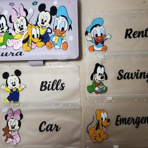 May include: A light purple binder featuring Disney characters and the name Laura, accompanied by five clear plastic zipper pouches. Each pouch displays a Disney character and a black label with the text Rent, Savings, Emergency, Bills, or Car.