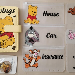 May include: A yellow A6 binder with a Winnie the Pooh graphic and the word Savings, accompanied by five translucent zipper pouches featuring Winnie the Pooh, Eeyore, Tigger, Roo, and Piglet with labels for House, Car, Insurance, and Emergency.