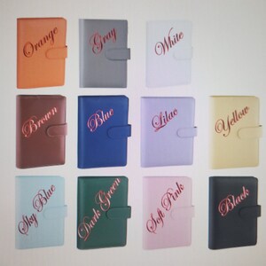 May include: A collection of eleven rectangular planners with textured covers and side closure tabs. Each planner features a color name in cursive script: Orange, Gray, White, Brown, Blue, Lilac, Yellow, Sky Blue, Dark Green, Soft Pink, and Black.