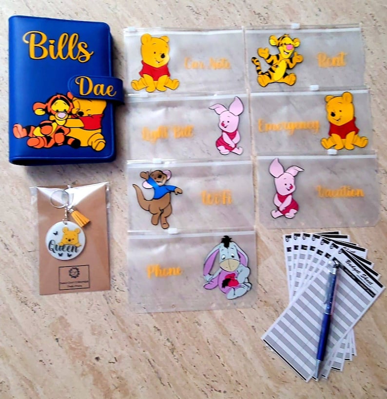 May include: A blue budget binder with Winnie the Pooh and Tigger graphics, labeled Bills Dae. Includes six translucent zipper pouches featuring various character illustrations and text labels, a circular keychain with a Pooh graphic, and a stack of budget sheets.