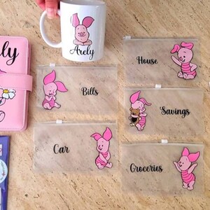 May include: A pink budget binder with the name Arely, a white ceramic mug with a Piglet graphic and the name Arely, five clear zippered pouches with Piglet illustrations and labels for House, Bills, Savings, Car, and Groceries, and a pink pen.