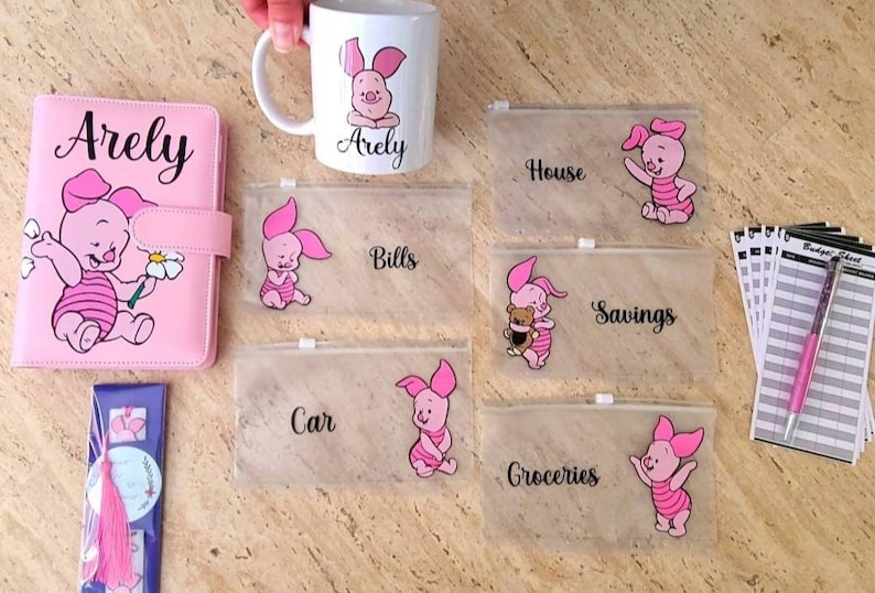 May include: A pink budget binder with the name Arely, a white ceramic mug with a Piglet graphic and the name Arely, five clear zippered pouches with Piglet illustrations and labels for House, Bills, Savings, Car, and Groceries, and a pink pen.