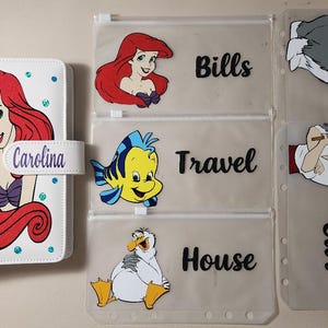 May include: A white binder featuring a red-haired mermaid character and the name Carolina, accompanied by five translucent zipper pouches. The pouches display cartoon characters and the labels Bills, Travel, House, and Car in black text.