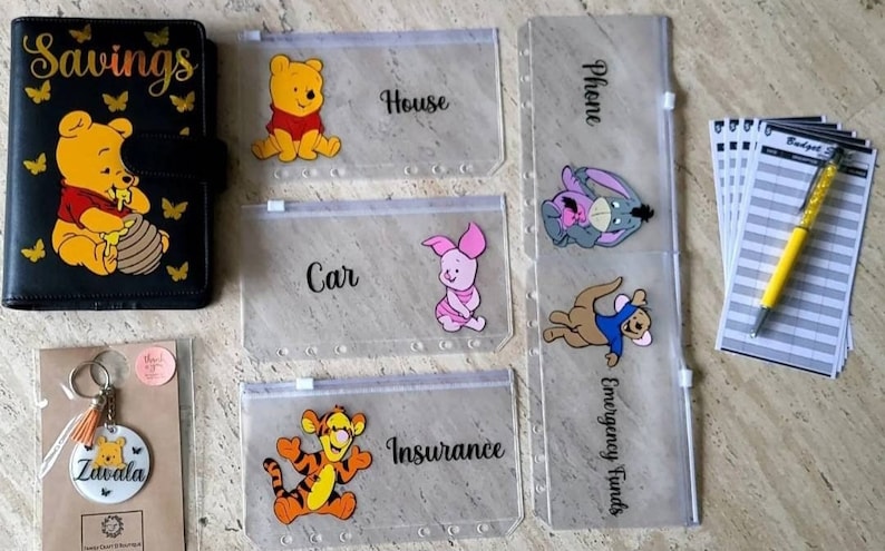 May include: A black savings binder featuring Winnie the Pooh, five clear zippered cash envelopes with character decals and labels like House, Car, and Insurance, a personalized circular keychain, a yellow pen, and a stack of budget sheets.