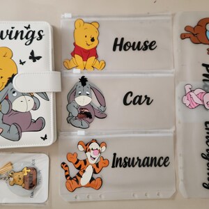 May include: A white A6 binder with a Winnie the Pooh and Eeyore graphic and the word Savings, three translucent zipper pouches labeled House, Car, and Insurance with character graphics, and a keychain with a Winnie the Pooh charm on a card.