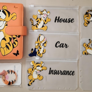 May include: An orange A6 binder featuring a Tigger graphic, accompanied by four translucent zipper pouches labeled House, Car, Insurance, and Emergency Phone, each decorated with Tigger illustrations, and a circular Tigger-themed keychain.