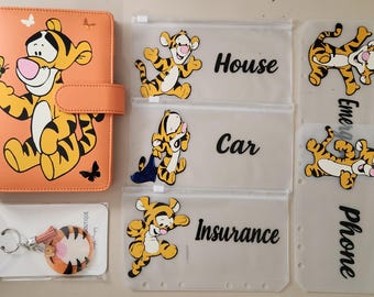 Op de afbeelding: Een oranje A6-map met een Tigger-afbeelding, vergezeld van vier doorzichtige ritszakjes met de labels House, Car, Insurance en Emergency Phone, elk versierd met Tigger-illustraties, en een ronde Tigger-sleutelhanger.