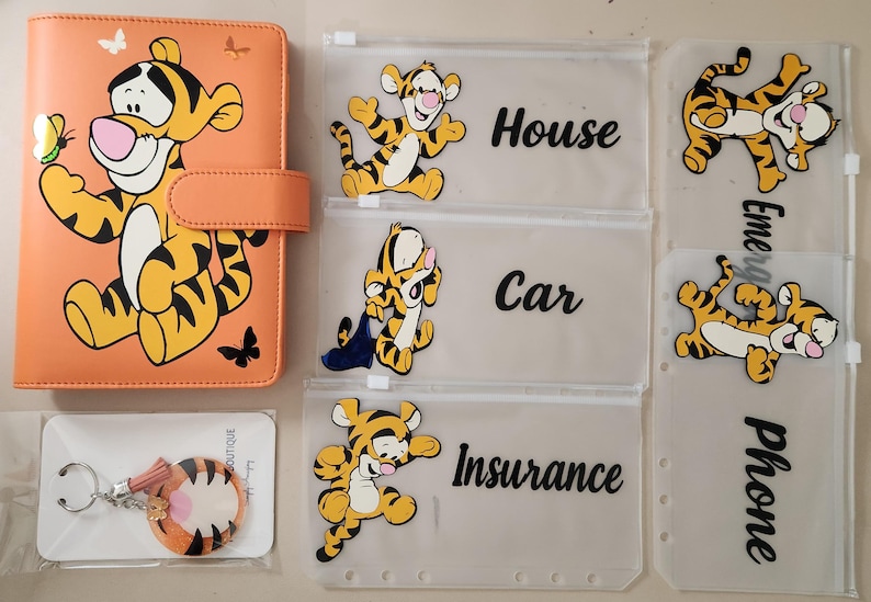 May include: An orange A6 binder featuring a Tigger graphic, accompanied by four translucent zipper pouches labeled House, Car, Insurance, and Emergency Phone, each decorated with Tigger illustrations, and a circular Tigger-themed keychain.