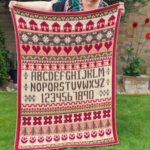 Puede incluir: Una manta de crochet roja, blanca y marrón con un patrón de casas, corazones, árboles, un alfabeto y números. La manta está siendo sostenida por una persona.