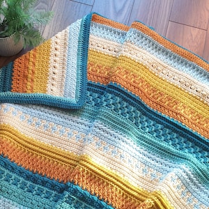 Puede incluir: Una manta de ganchillo con rayas de color azul verdoso, azul, amarillo y naranja. La manta está doblada por la mitad y tiene un borde de color naranja y azul verdoso.