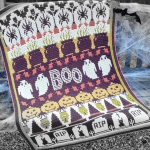 Puede incluir: Una manta de ganchillo de Halloween con un esquema de colores morado, naranja, blanco y verde. La manta presenta una variedad de dise&ntilde;os de tem&aacute;tica de Halloween, incluyendo fantasmas, calabazas, murci&eacute;lagos y l&aacute;pidas. La manta est&aacute; escrita "BOO" en el centro.
