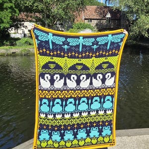Puede incluir: Una colorida manta de ganchillo con un patrón repetido de libélulas, cisnes y ranas. La manta tiene un fondo azul marino con detalles en turquesa, amarillo y blanco. La manta está bordeada con un borde amarillo.