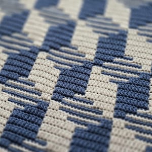 Op de afbeelding: Close-up van een gehaakte deken met een geometrisch patroon. Het ontwerp heeft afwisselende rijen blauw en gebroken wit, waardoor een getextureerd zigzag-effect ontstaat. De deken is gemaakt van zacht garen.