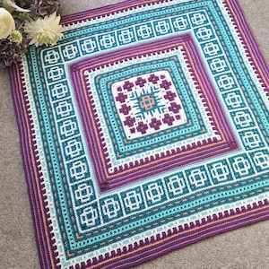 Op de afbeelding: Een vierkant, handgehaakt kleed met een geometrisch ontwerp. Het kleed heeft een centraal vierkant met een bloemenpatroon, omgeven door concentrische banden van turquoise, paars en wit. De rand is dieppaars.