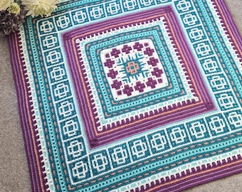 Op de afbeelding: Een vierkant, handgehaakt kleed met een geometrisch ontwerp. Het kleed heeft een centraal vierkant met een bloemenpatroon, omgeven door concentrische banden van turquoise, paars en wit. De rand is dieppaars.