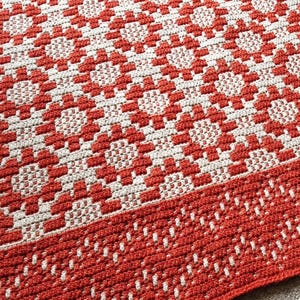 Op de afbeelding: Een rood en wit gehaakt tapijt met een geometrisch patroon. Het tapijt heeft een franje rand.