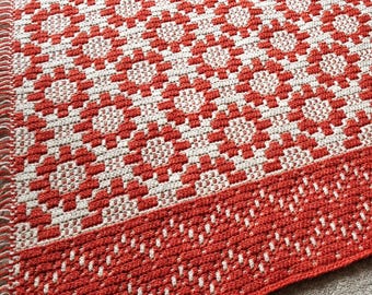 Puede incluir: Una alfombra de crochet roja y blanca con un patrón geométrico. La alfombra tiene un borde de flecos.