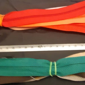 May include: A bundle of red, orange, and brown zippers with a rubber band holding them together. Below is a bundle of green, brown, and beige zippers with a rubber band holding them together.