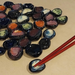 Puede incluir: Una colección de pequeños reposa palillos de cerámica en varios colores, incluyendo azul, naranja, morado y verde. Los reposa palillos están dispuestos sobre una superficie de color claro, con un par de palillos rojos apoyados en uno. Acabado brillante.