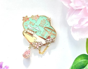 May include: A gold-colored enamel pin with a light blue background and the words "YOU NEVER WALK ALONE". The pin features a beach scene with cherry blossom accents and a dangling pink charm. The pin is decorated with gold-colored chain.