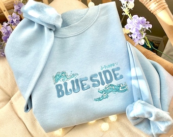 May include: A light blue sweatshirt with the word "BLUESIDE" embroidered in a darker blue, along with Japanese text and wave designs. The sweatshirt is folded on a beige cushion, with decorative lights and purple flowers in the background.