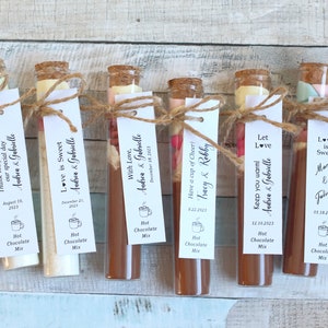 May include: Ten small glass tubes filled with hot chocolate mix and topped with marshmallows and other toppings. Each tube has a small tag tied around it with a personalized message and the date of the event. The tags read "Thank you for coming Andrea & Gabrielle", "Thank you for sharing our special day Andrea & Gabrielle", "Love is Sweet Andrea & Gabrielle", "With Love, Andrea & Gabrielle", "Have a cup of Cheer! Tracy & Robby", "Let Love Keep you warm Andrea & Gabrielle", "Love is Sweet Mary & Jolien", "Love is Sweet Andrea & Gabrielle".