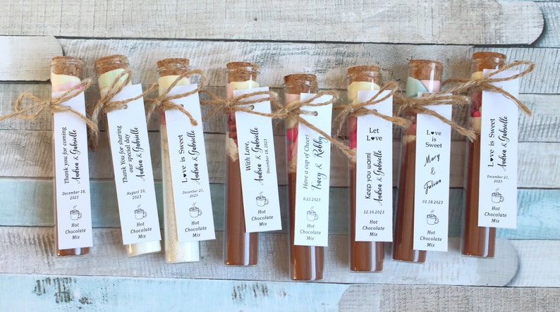 May include: Ten small glass tubes filled with hot chocolate mix and topped with marshmallows and other toppings. Each tube has a small tag tied around it with a personalized message and the date of the event. The tags read "Thank you for coming Andrea & Gabrielle", "Thank you for sharing our special day Andrea & Gabrielle", "Love is Sweet Andrea & Gabrielle", "With Love, Andrea & Gabrielle", "Have a cup of Cheer! Tracy & Robby", "Let Love Keep you warm Andrea & Gabrielle", "Love is Sweet Mary & Jolien", "Love is Sweet Andrea & Gabrielle".