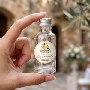 May include: A small, clear glass bottle with a silver screw cap, featuring a circular label with a lemon illustration and the text Limoncello with love, Hazel & Lincoln, and the date 12.12.2027 printed in black and gold script fonts.