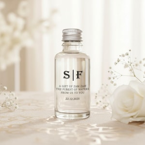Pode incluir: Uma garrafa de vidro transparente com tampa prateada, com as iniciais "S|F" em preto. A garrafa tem o texto "A GIFT OF ZAM ZAM THE PUREST OF WATERS FROM US TO YOU 22.12.2025". A garrafa está sobre uma superfície clara.