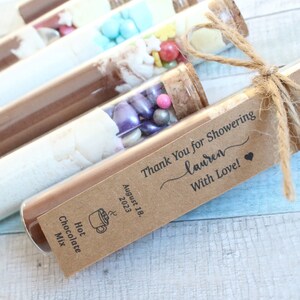 May include: A set of glass test tubes filled with various ingredients, including chocolate powder, marshmallows, and sprinkles. The tubes are tied with twine and a brown paper tag that reads "Thank You for Showering Lauren With Love! August 18, 2023".