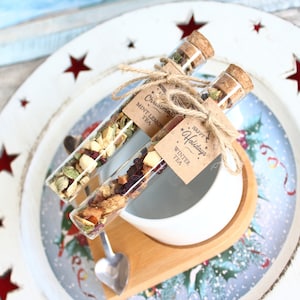 May include: Two glass tubes filled with tea, labeled "Christmas Mint Lemon Tea" and "Happy Holidays Winter Tea", are tied with twine. They rest on a wooden tray with a white bowl and a decorative plate with red star cutouts.