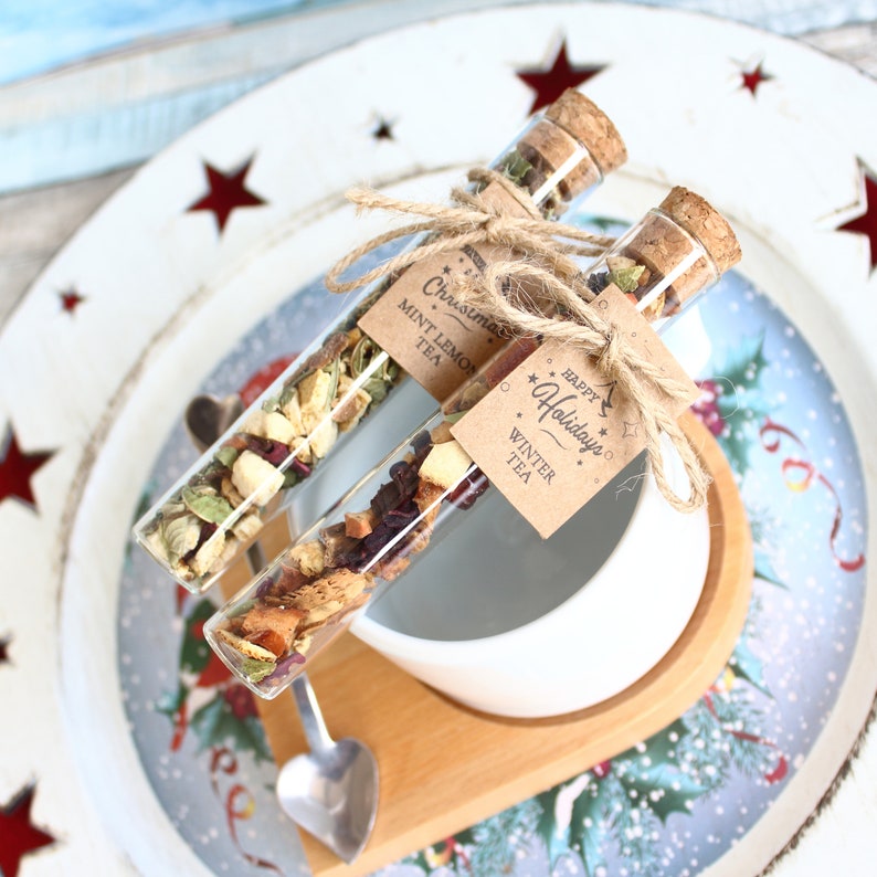May include: Two glass tubes filled with tea, labeled "Christmas Mint Lemon Tea" and "Happy Holidays Winter Tea", are tied with twine. They rest on a wooden tray with a white bowl and a decorative plate with red star cutouts.
