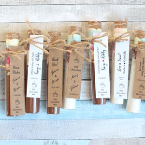 May include: A collection of small glass test tubes filled with hot chocolate mix and topped with marshmallows. Each tube has a personalized tag with a message and the names of the couple. The tags are tied with twine and a small heart.