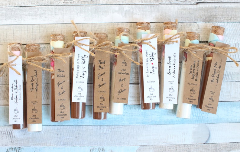 May include: A collection of small glass test tubes filled with hot chocolate mix and topped with marshmallows. Each tube has a personalized tag with a message and the names of the couple. The tags are tied with twine and a small heart.