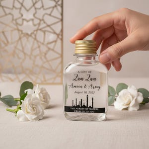Pode incluir: Uma garrafa de vidro transparente com tampa dourada, com o texto "A GIFT OF Zam Zam Ameen & Arooj August 18, 2025". A garrafa tem um rótulo preto com uma silhueta de edifícios e o texto "THE PUREST OF WATERS FROM US TO YOU."